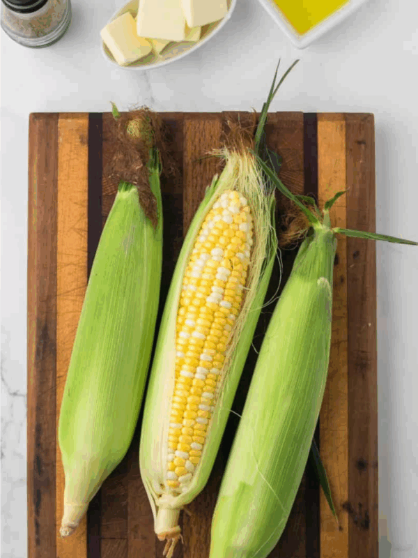Three ears of corn, one partially husked, rest on a wooden cutting board. Nearby are salt and pepper shakers, a dish of butter, and a small bowl of oil—perfect for learning how to preserve corn at home.