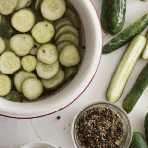 Sliced cucumbers soaking in water, whole cucumbers, pickling spices, and salt are arranged on a white surface. A green label reads “blessthismessplease.com.” Learn how to preserve cucumber with these simple ingredients.