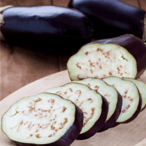 A close-up of sliced eggplant on a wooden cutting board, with two whole eggplants in the background. The website blessthismessplease.com is printed on the bottom edge, perfect for illustrating how to preserve eggplant.