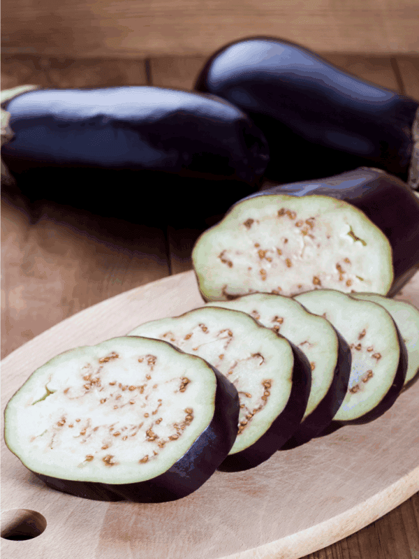A close-up of sliced eggplant on a wooden cutting board, with two whole eggplants in the background. The website blessthismessplease.com is printed on the bottom edge, perfect for illustrating how to preserve eggplant.