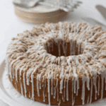 A round peach coffee cake with crumb topping and white icing drizzled on top, displayed on a white plate. Stacked plates and forks are visible in the blurred background.