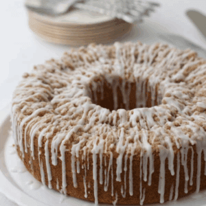 A round peach coffee cake with crumb topping and white icing drizzled on top, displayed on a white plate. Stacked plates and forks are visible in the blurred background.