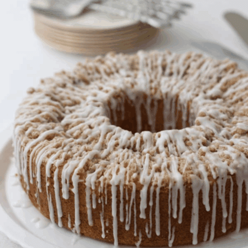 A round peach coffee cake with crumb topping and white icing drizzled on top, displayed on a white plate. Stacked plates and forks are visible in the blurred background.
