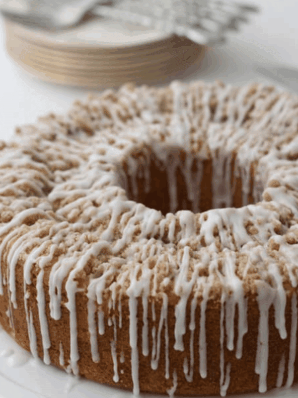A round peach coffee cake with crumb topping and white icing drizzled on top, displayed on a white plate. Stacked plates and forks are visible in the blurred background.