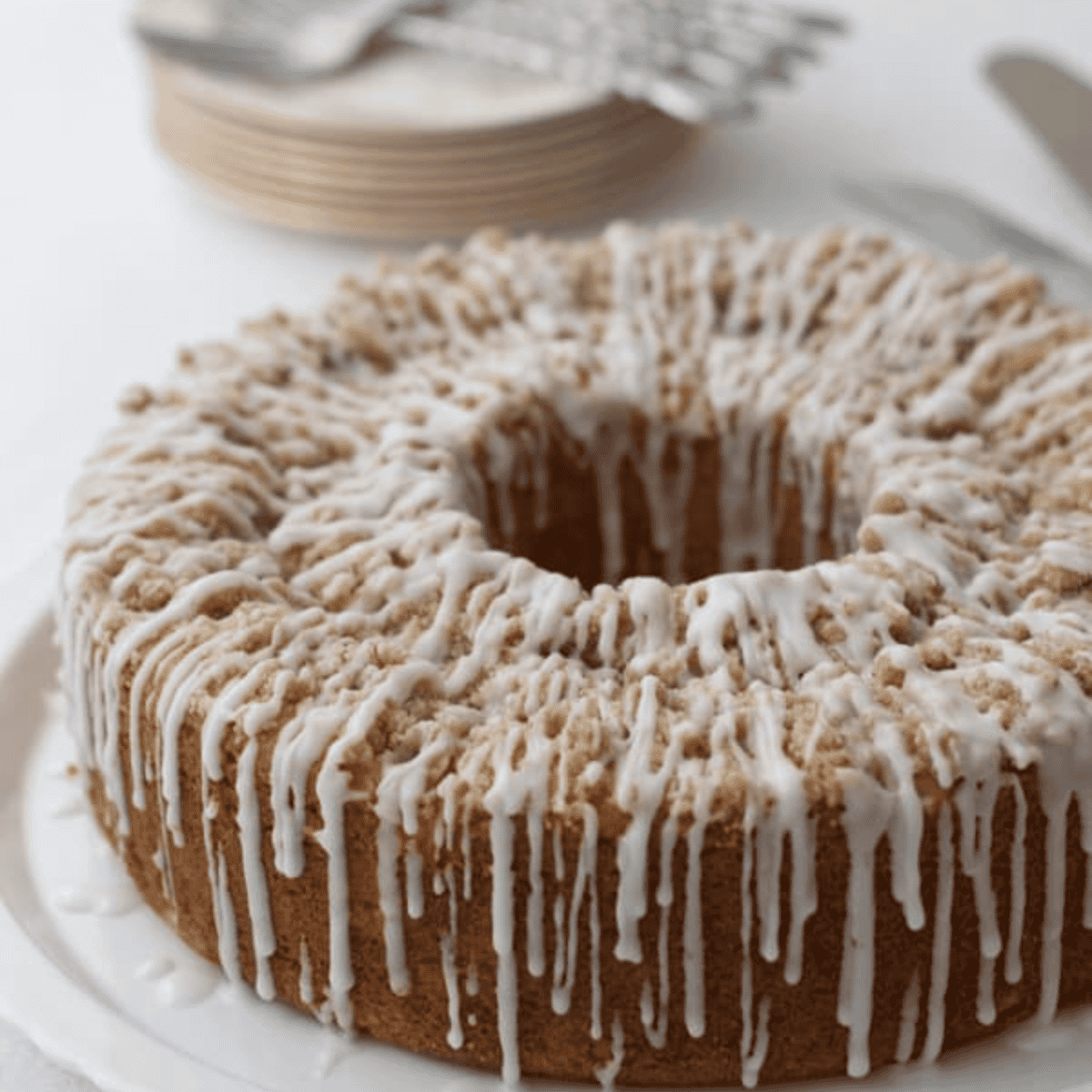 A round peach coffee cake with crumb topping and white icing drizzled on top, displayed on a white plate. Stacked plates and forks are visible in the blurred background.