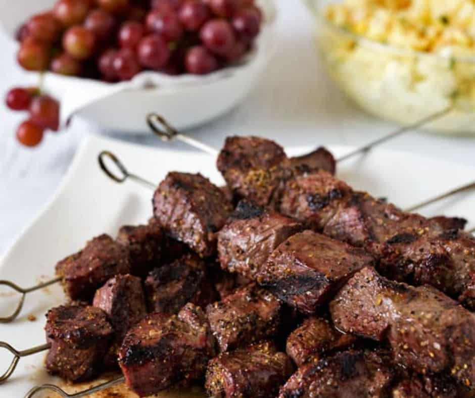 Grilled beef kebabs on skewers are served on a white plate. In the background, a bowl of red grapes and another bowl with a yellow, likely potato-based side dish complete this tempting beef kebabs recipe scene.