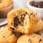 A close-up of chocolate chip muffins, with one muffin showing a bite taken out, revealing gooey melted chocolate chunks inside. Other whole muffins and a bowl of chocolate chips are in the background featuring a chocolate chip muffin recipe.