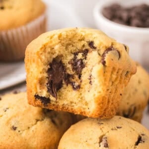 A close-up of chocolate chip muffins, with one muffin showing a bite taken out, revealing gooey melted chocolate chunks inside. Other whole muffins and a bowl of chocolate chips are in the background featuring a chocolate chip muffin recipe.