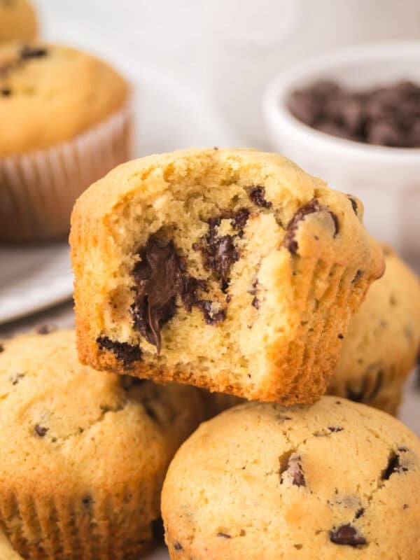 A close-up of chocolate chip muffins, with one muffin showing a bite taken out, revealing gooey melted chocolate chunks inside. Other whole muffins and a bowl of chocolate chips are in the background featuring a chocolate chip muffin recipe.