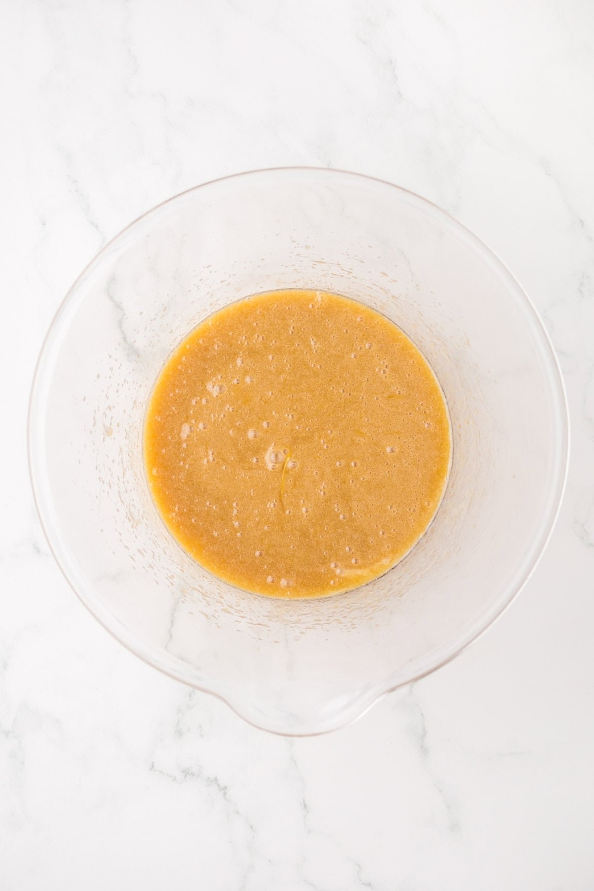A clear glass mixing bowl with a light brown, slightly frothy liquid batter sits on a white marble surface.