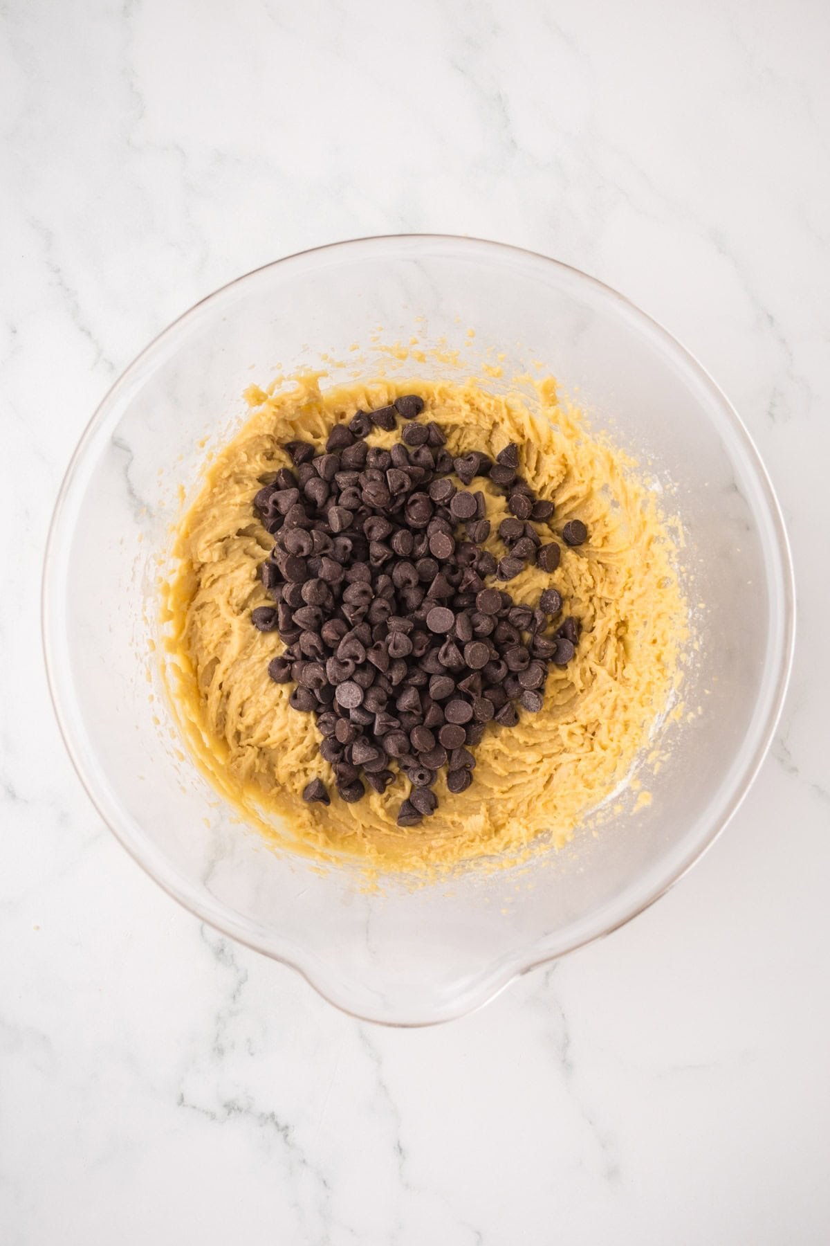 A glass mixing bowl with cookie dough and a heap of chocolate chips on top, sitting on a white marble surface.
