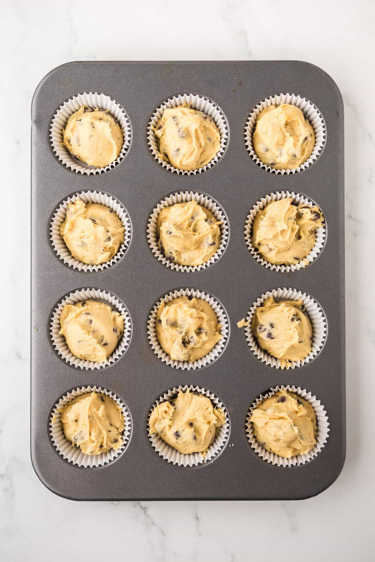 A muffin tin with 12 paper liners filled with chocolate chip cookie dough, ready to be baked, sits on a white marble surface.