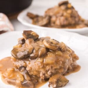 A chopped steak patty topped with brown mushroom and onion gravy is served on a white plate, with another plate of the same dish and a skillet blurred in the background.