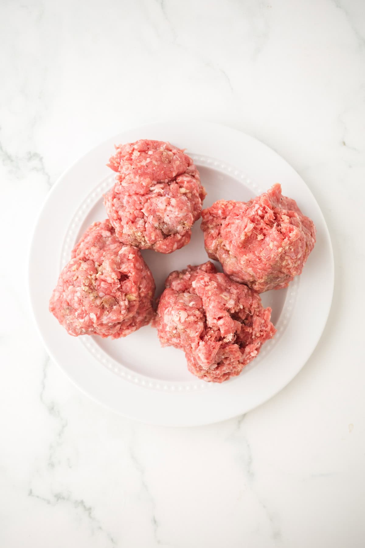 Four raw ground beef portions, perfect for making chopped steak, are shaped into rough balls and placed on a white plate, viewed from above on a white marble surface.