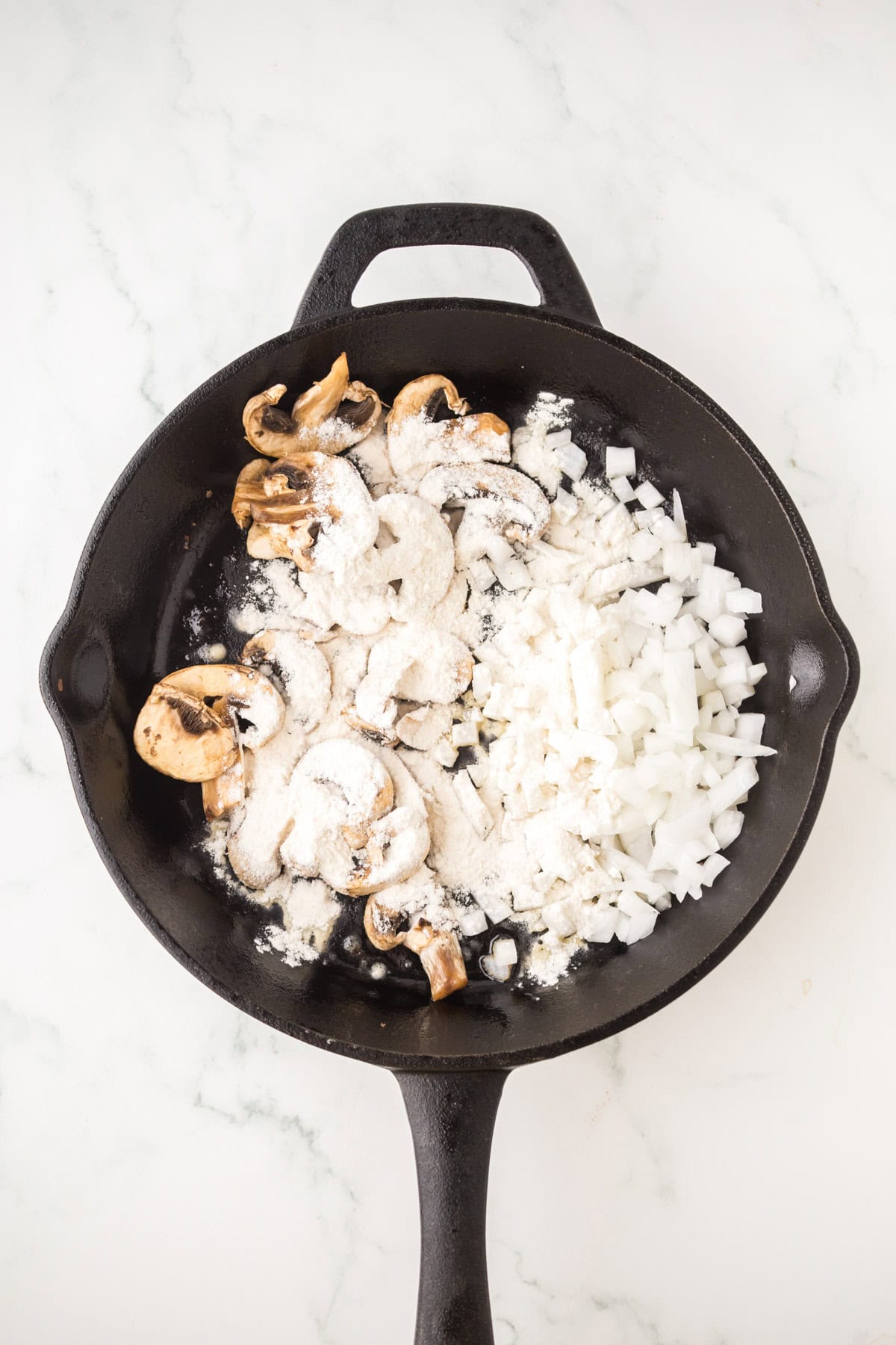 A cast iron skillet with sliced mushrooms, chopped onions, flour, and chopped steak on a white marble surface, ready to be cooked.