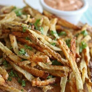 A pile of crispy, seasoned garlic parmesan French fries garnished with chopped parsley on a wooden board, with a small bowl of creamy dipping sauce in the background.