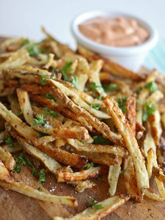 A pile of crispy, seasoned garlic parmesan French fries garnished with chopped parsley on a wooden board, with a small bowl of creamy dipping sauce in the background.