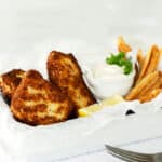 A white tray with crispy halibut fish and chips, golden French fries, lemon wedges, and a small bowl of tartar sauce garnished with parsley. A fork and knife rest on a white textured surface beside the tray.