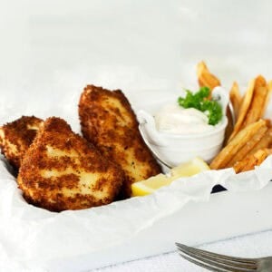 A white tray with crispy halibut fish and chips, golden French fries, lemon wedges, and a small bowl of tartar sauce garnished with parsley. A fork and knife rest on a white textured surface beside the tray.