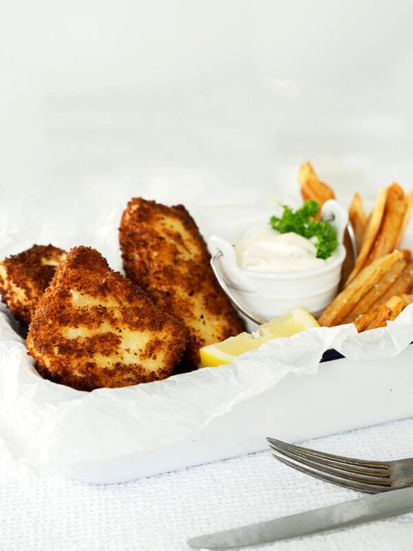 A white tray with crispy halibut fish and chips, golden French fries, lemon wedges, and a small bowl of tartar sauce garnished with parsley. A fork and knife rest on a white textured surface beside the tray.