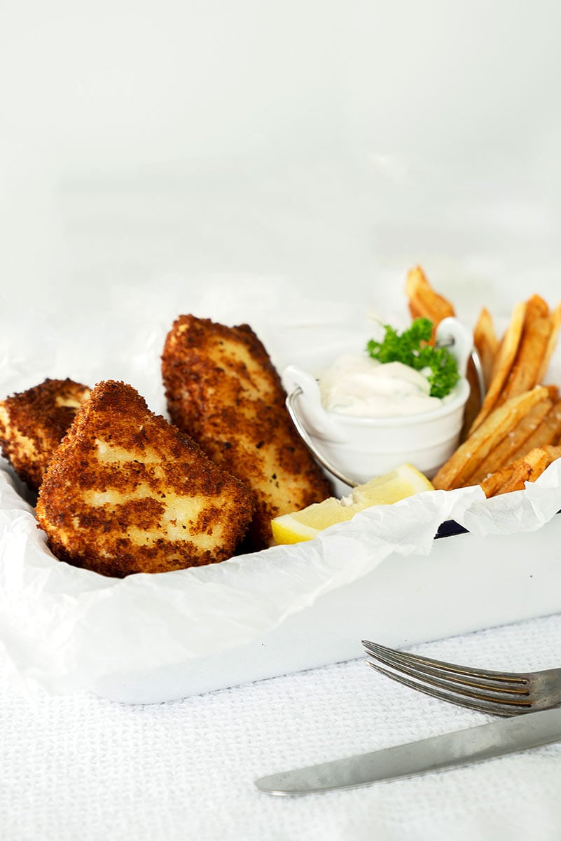 A white tray with crispy halibut fish and chips, golden French fries, lemon wedges, and a small bowl of tartar sauce garnished with parsley. A fork and knife rest on a white textured surface beside the tray.