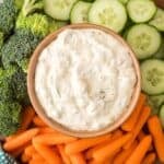 A bowl of creamy Knorr veggie spinach dip sits surrounded by fresh broccoli florets, sliced cucumbers, and baby carrots, all artfully arranged on a platter.