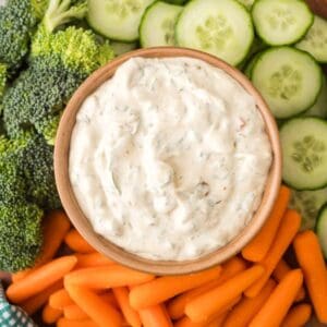 A bowl of creamy Knorr veggie spinach dip sits surrounded by fresh broccoli florets, sliced cucumbers, and baby carrots, all artfully arranged on a platter.