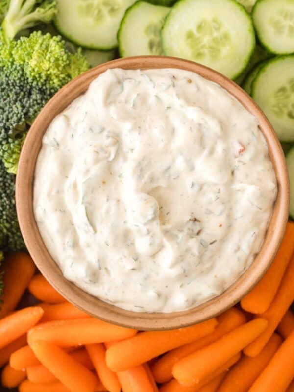 A bowl of creamy Knorr veggie spinach dip sits surrounded by fresh broccoli florets, sliced cucumbers, and baby carrots, all artfully arranged on a platter.