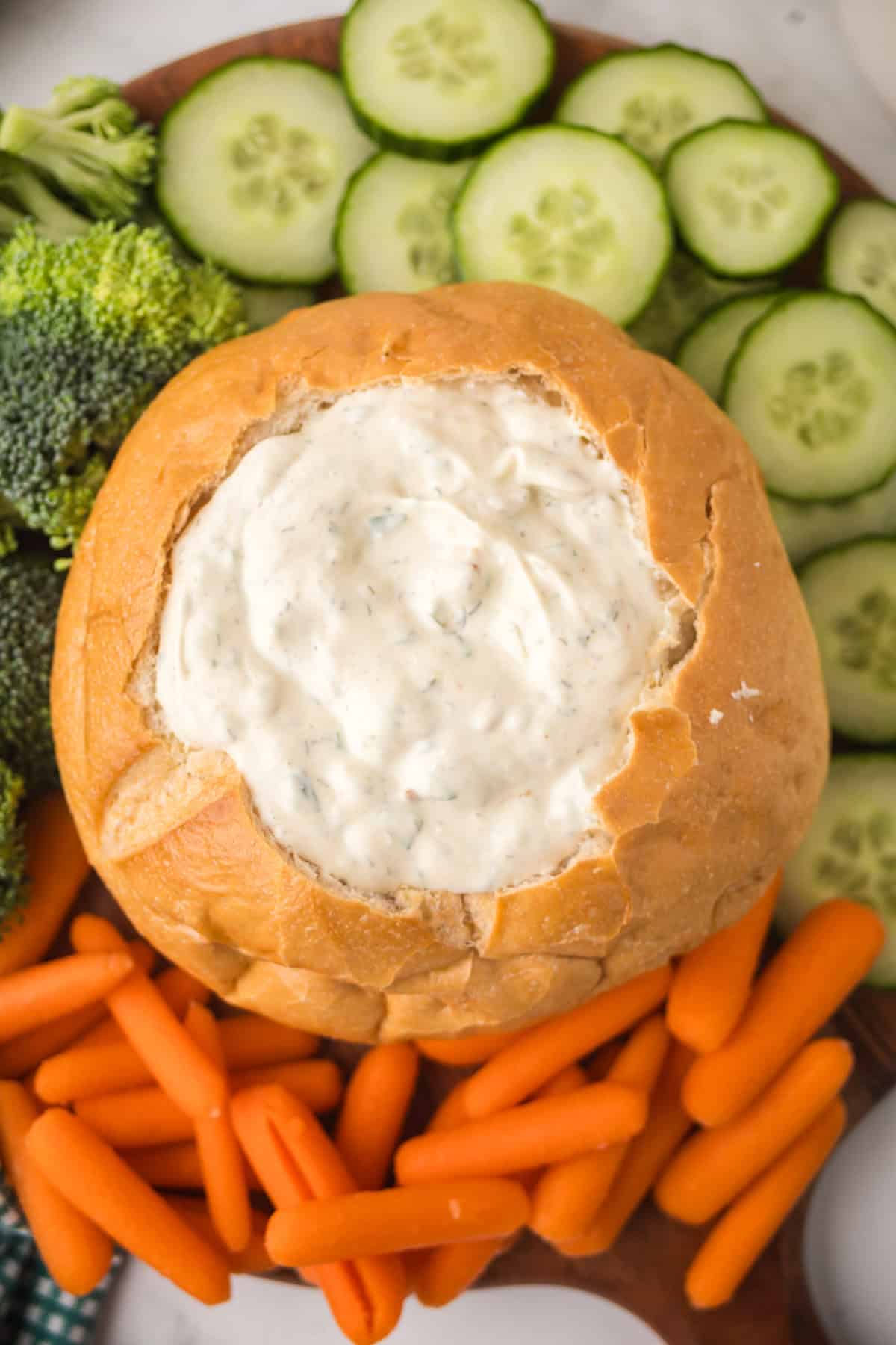 A round bread loaf hollowed out and filled with creamy Knorr veggie spinach dip is surrounded by fresh cucumber slices, broccoli florets, and baby carrots on a wooden serving board.