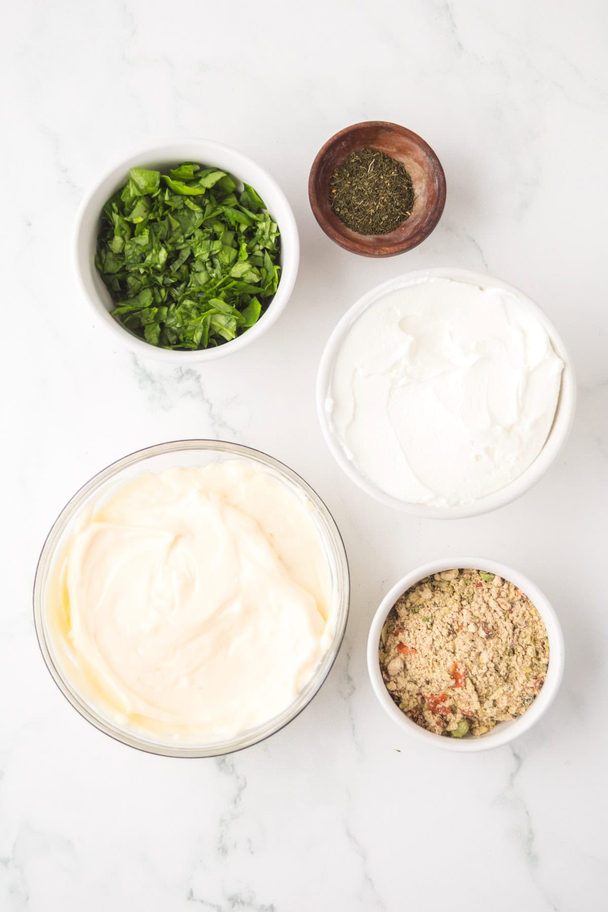 Five bowls arranged on a white marble surface, containing ingredients for knorr veggie spinach dip: chopped fresh herbs, dried herbs, creamy white cheese, mayonnaise, and seasoned breadcrumbs.