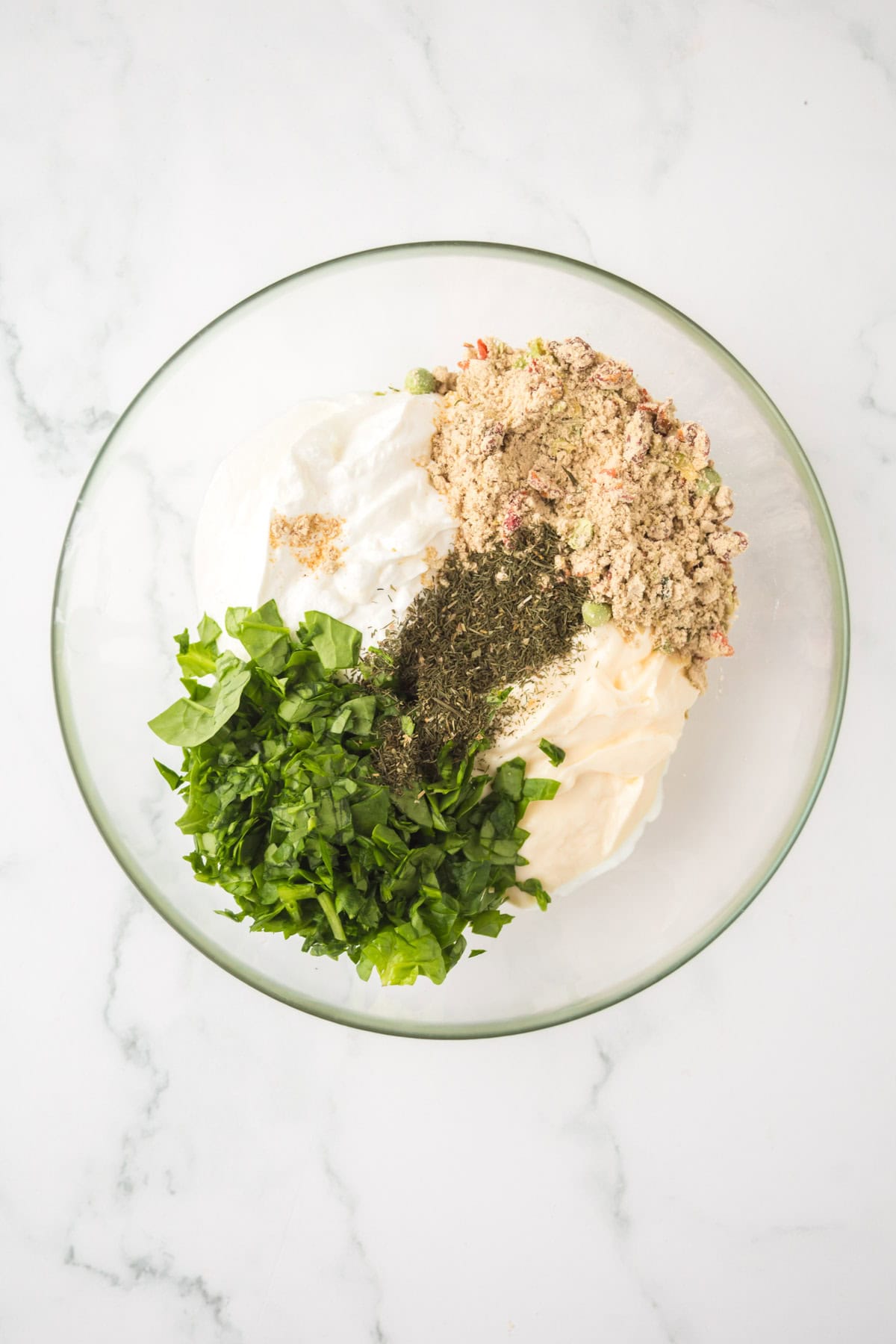 A glass bowl on a marble surface holds the separated ingredients for a classic Knorr veggie spinach dip—chopped spinach, dried herbs, mayonnaise, sour cream, and dry soup mix—all ready to be blended together.