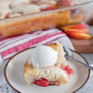 A slice of peach raspberry cobbler and strawberry cobbler topped with vanilla ice cream sits on a plate with a spoon. In the background, a glass baking dish with more cobbler and fresh peach slices are visible.