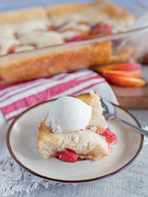 A slice of peach raspberry cobbler and strawberry cobbler topped with vanilla ice cream sits on a plate with a spoon. In the background, a glass baking dish with more cobbler and fresh peach slices are visible.
