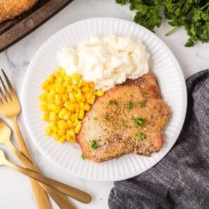 A white plate with shake and bake pork chops garnished with herbs, a serving of mashed potatoes, and yellow corn. Gold utensils are on the side, and fresh parsley is partly visible at the top right.