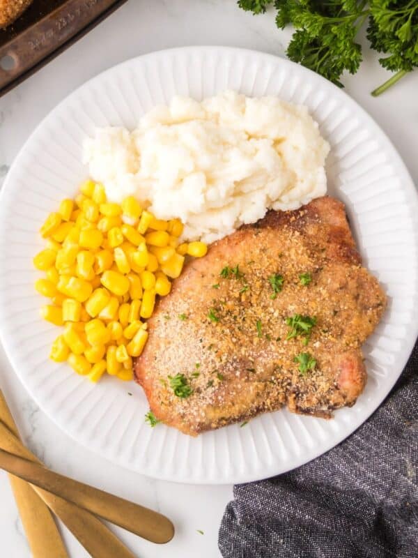 A white plate with shake and bake pork chops garnished with herbs, a serving of mashed potatoes, and yellow corn. Gold utensils are on the side, and fresh parsley is partly visible at the top right.