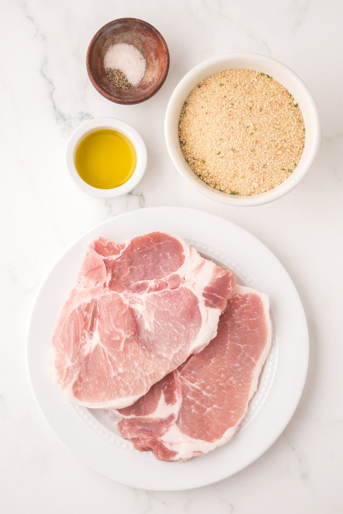 A white plate with shake and bake pork chops sits beside a bowl of breadcrumbs, a small dish of olive oil, and a small bowl containing salt and pepper on a white marble surface.
