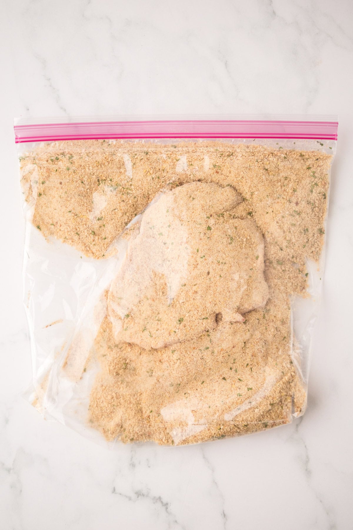 A raw chicken breast coated in seasoned breadcrumbs, similar to shake and bake pork chops, sits inside a sealed plastic zip-top bag on a white marble surface.