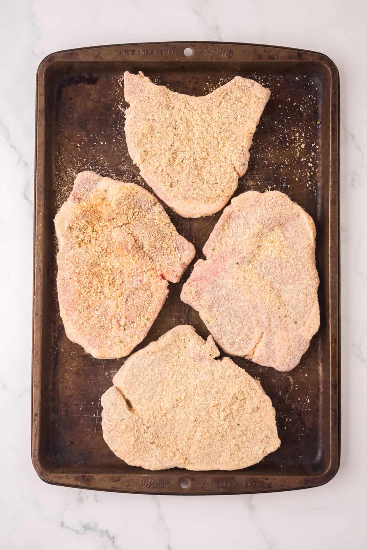 Four raw shake and bake pork chops are arranged on a dark baking sheet, ready to be cooked. The breading is evenly coated, and the pan rests on a white marble surface.
