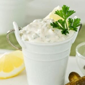 A small white pail filled with creamy tartar sauce, made from a classic tartar sauce recipe and garnished with parsley and a lemon wedge, sits on a white surface next to a pickle and a lemon slice.