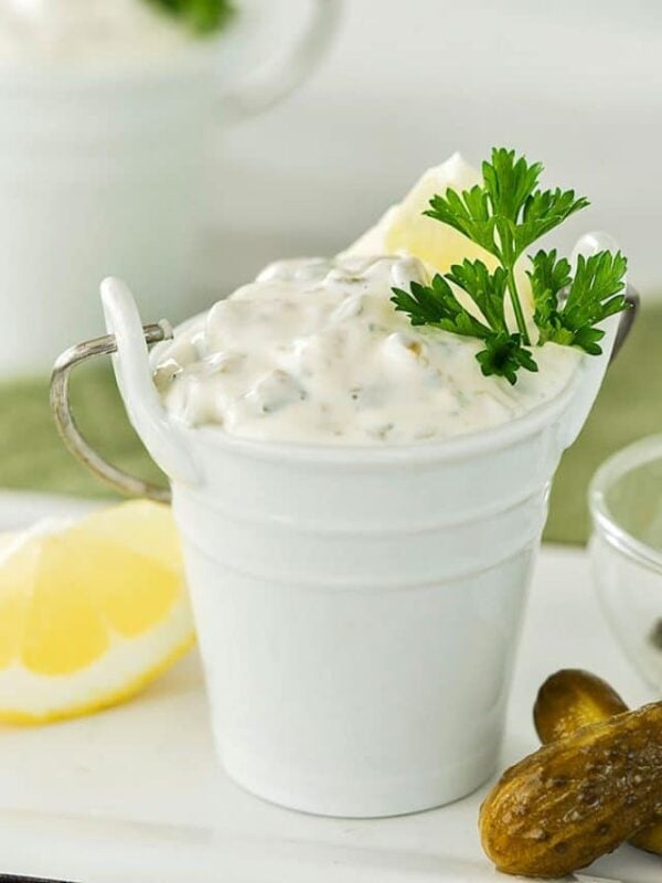 A small white pail filled with creamy tartar sauce, made from a classic tartar sauce recipe and garnished with parsley and a lemon wedge, sits on a white surface next to a pickle and a lemon slice.