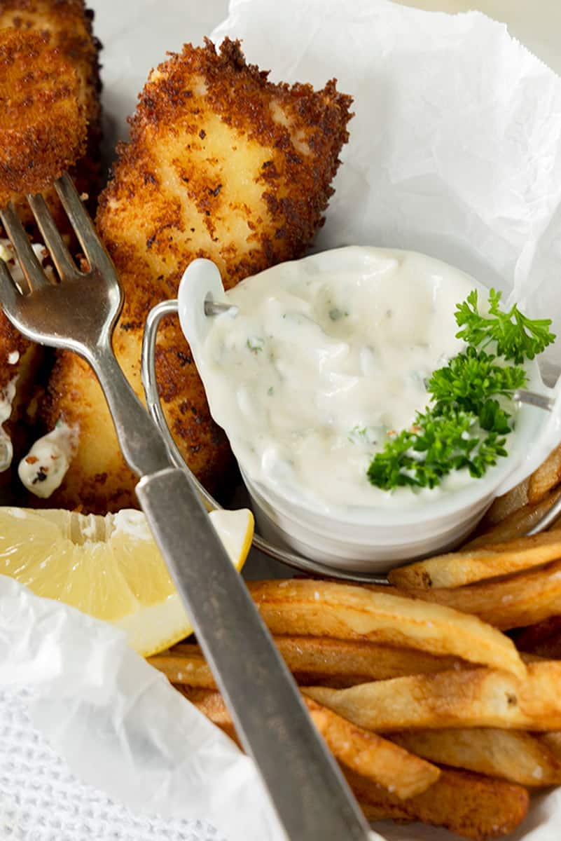 Close-up of crispy fish fillets with French fries, a wedge of lemon, and a small bowl of creamy tartar sauce recipe garnished with parsley on parchment paper, with a fork resting beside the food.