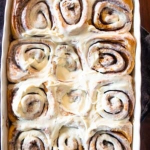 A white baking dish filled with freshly baked gingerbread cinnamon rolls topped with creamy white icing, sitting on a wooden surface with a dark cloth underneath. The rolls are golden brown and swirled.