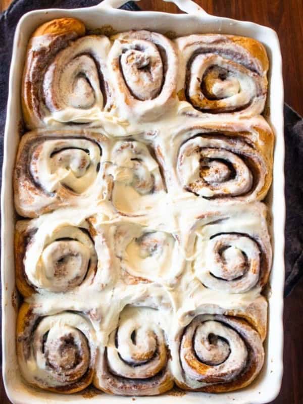 A white baking dish filled with freshly baked gingerbread cinnamon rolls topped with creamy white icing, sitting on a wooden surface with a dark cloth underneath. The rolls are golden brown and swirled.