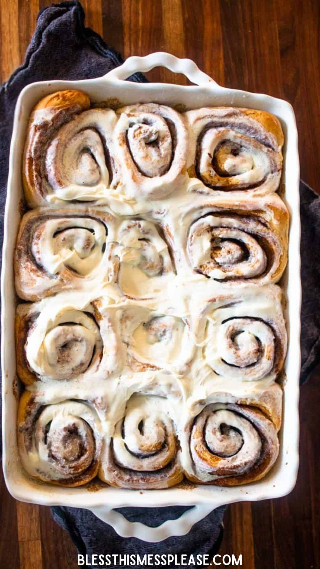 A white baking dish filled with freshly baked gingerbread cinnamon rolls topped with creamy white icing, sitting on a wooden surface with a dark cloth underneath. The rolls are golden brown and swirled.