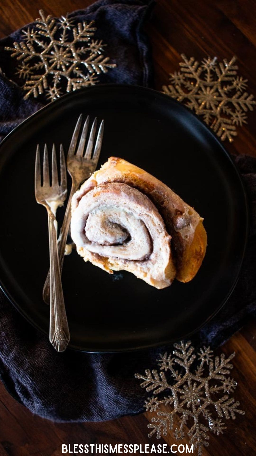 A frosted gingerbread cinnamon roll on a black plate with two forks, set on a dark cloth. Decorative gold snowflakes and the website blessthismessplease.com are also visible.