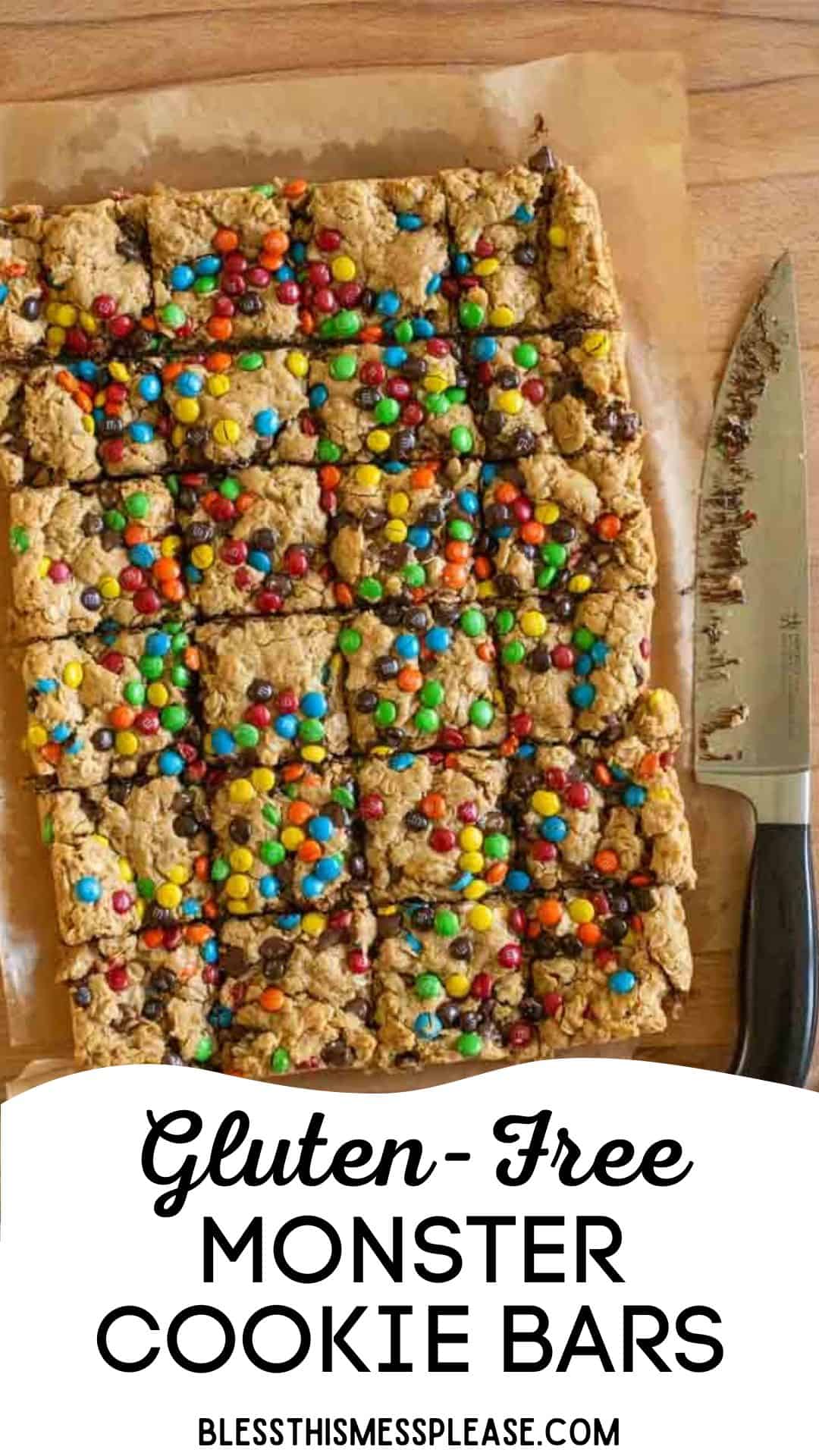 A tray of freshly baked monster cookie bars, gluten-free and topped with colorful candy pieces, is sliced into squares on parchment paper. A large knife rests beside them. Text at the bottom reads Gluten-Free Monster Cookie Bars.