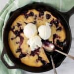 A skillet cherry dessert inspired by a cherry cobbler recipe, topped with three scoops of vanilla ice cream. A slice is removed, and two spoons rest on the skillet, which sits atop a green towel on a marble surface.