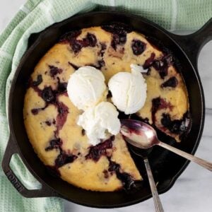 A skillet cherry dessert inspired by a cherry cobbler recipe, topped with three scoops of vanilla ice cream. A slice is removed, and two spoons rest on the skillet, which sits atop a green towel on a marble surface.