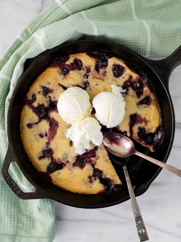 A skillet cherry dessert inspired by a cherry cobbler recipe, topped with three scoops of vanilla ice cream. A slice is removed, and two spoons rest on the skillet, which sits atop a green towel on a marble surface.