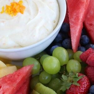 A bowl of dip made from a fruit dip recipe garnished with orange zest is surrounded by fresh fruit, including watermelon slices, green grapes, blueberries, raspberries, strawberries, kiwi, and pineapple.